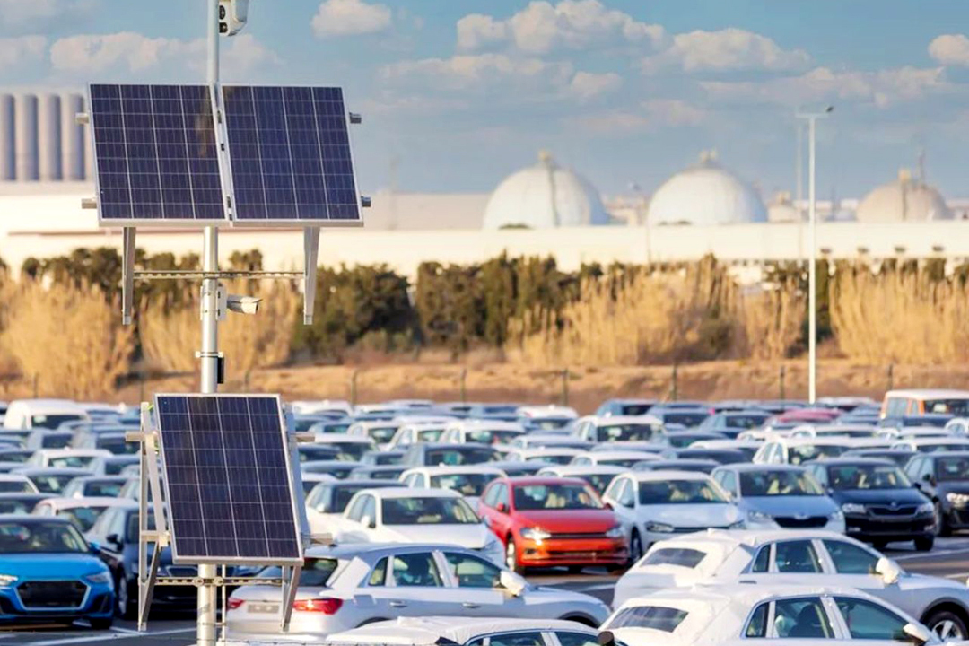 #PraCegoVer Na imagem podemos ver carros em um estacionamento onde vemos placas solares em um poste no alto. Ao fundo temos o céu azul e um campo com muitas árvores.
